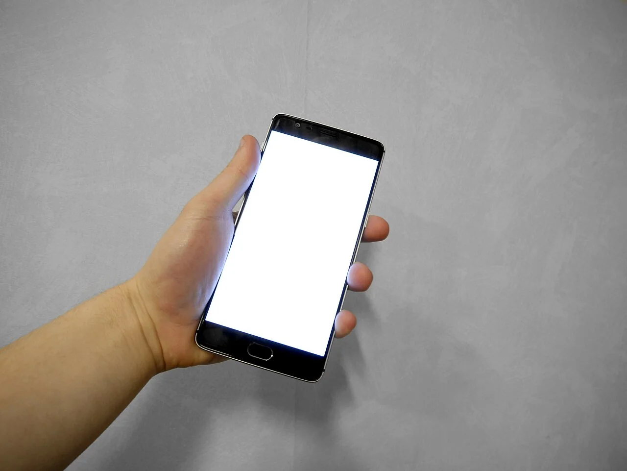 A hand holds a smartphone with a blank white screen against a grey wall.